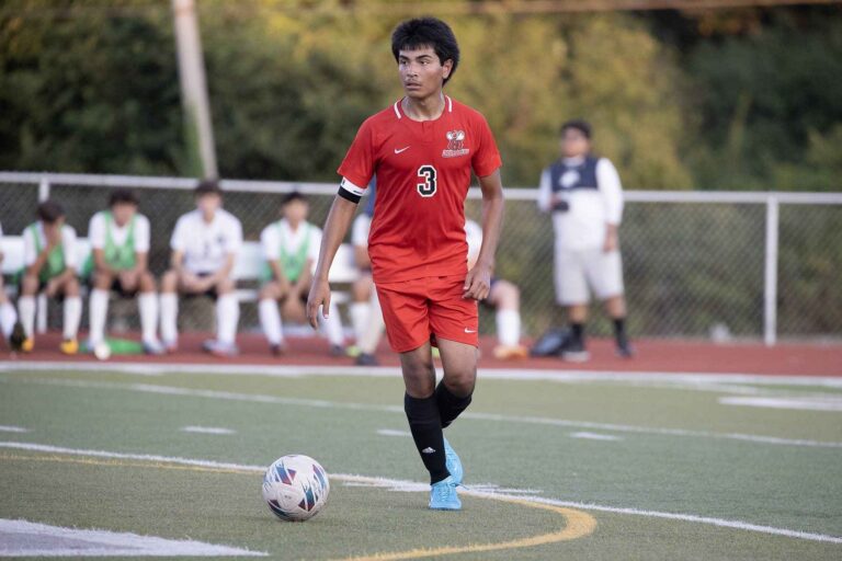 A soccer player wearing a red uniform stands on the field, ready to play, with green grass and goalposts in the background.