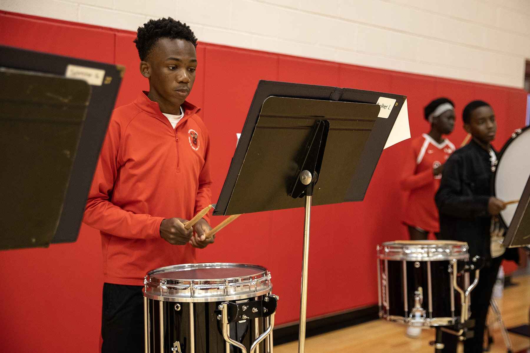 A young man wearing a red shirt energetically plays drums in a gym setting, surrounded by exercise equipment.