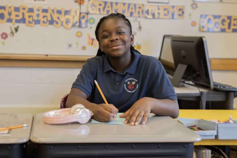 A young student sits at a desk, focused on writing with a pencil on a sheet of paper in front of her.