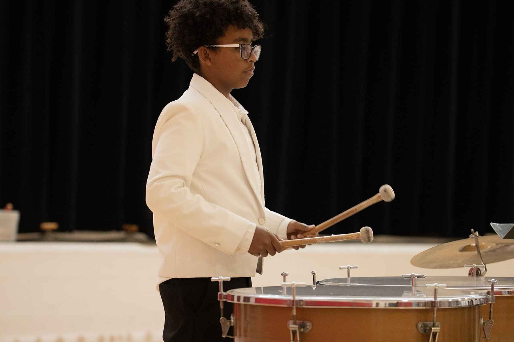 A young boy in a white suit energetically playing drums on stage, showcasing his musical talent and enthusiasm.