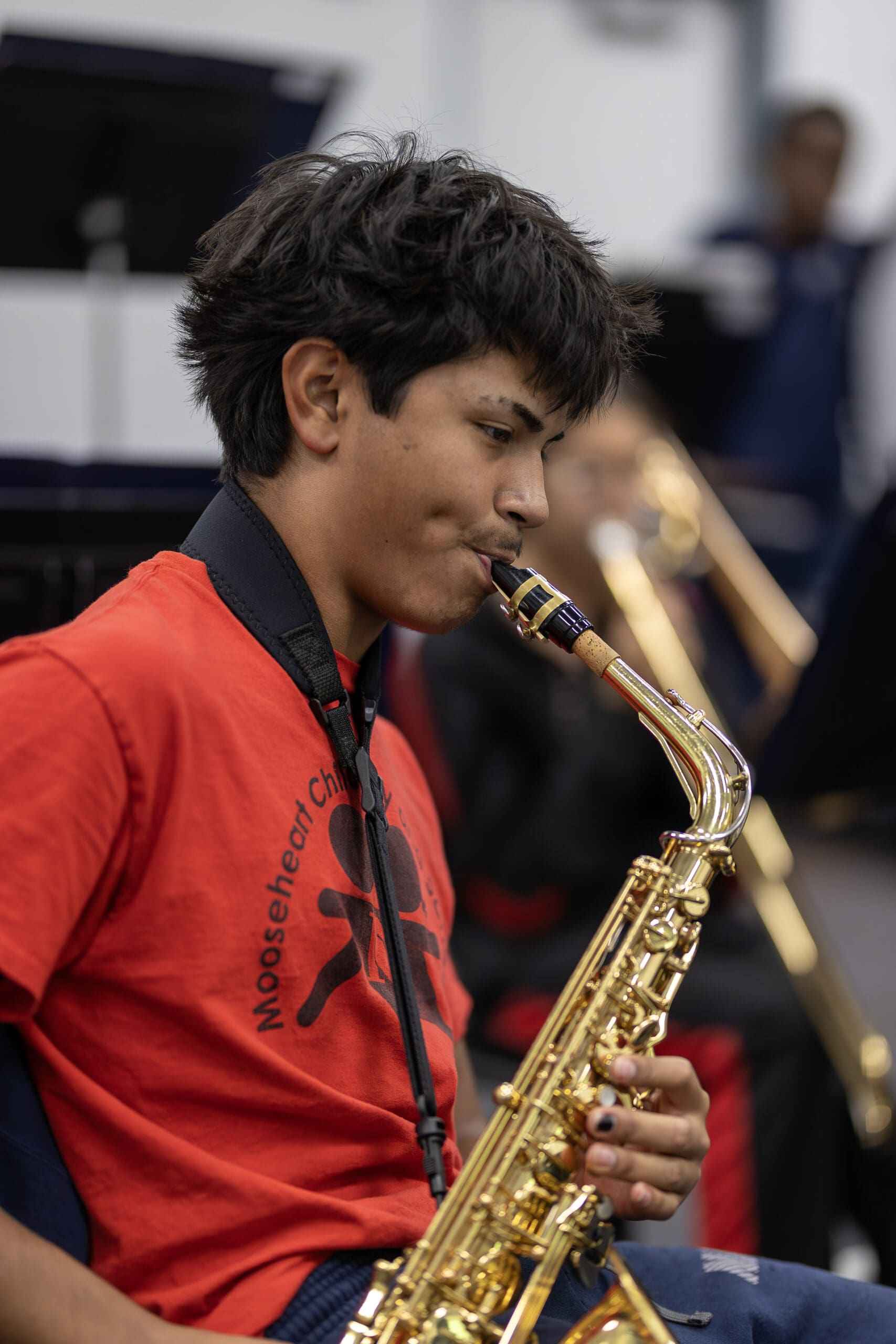 A boy is sitting down in the school gym, saxophone in hand and playing it, he looks focused as he plays this beautiful, gold instrument.