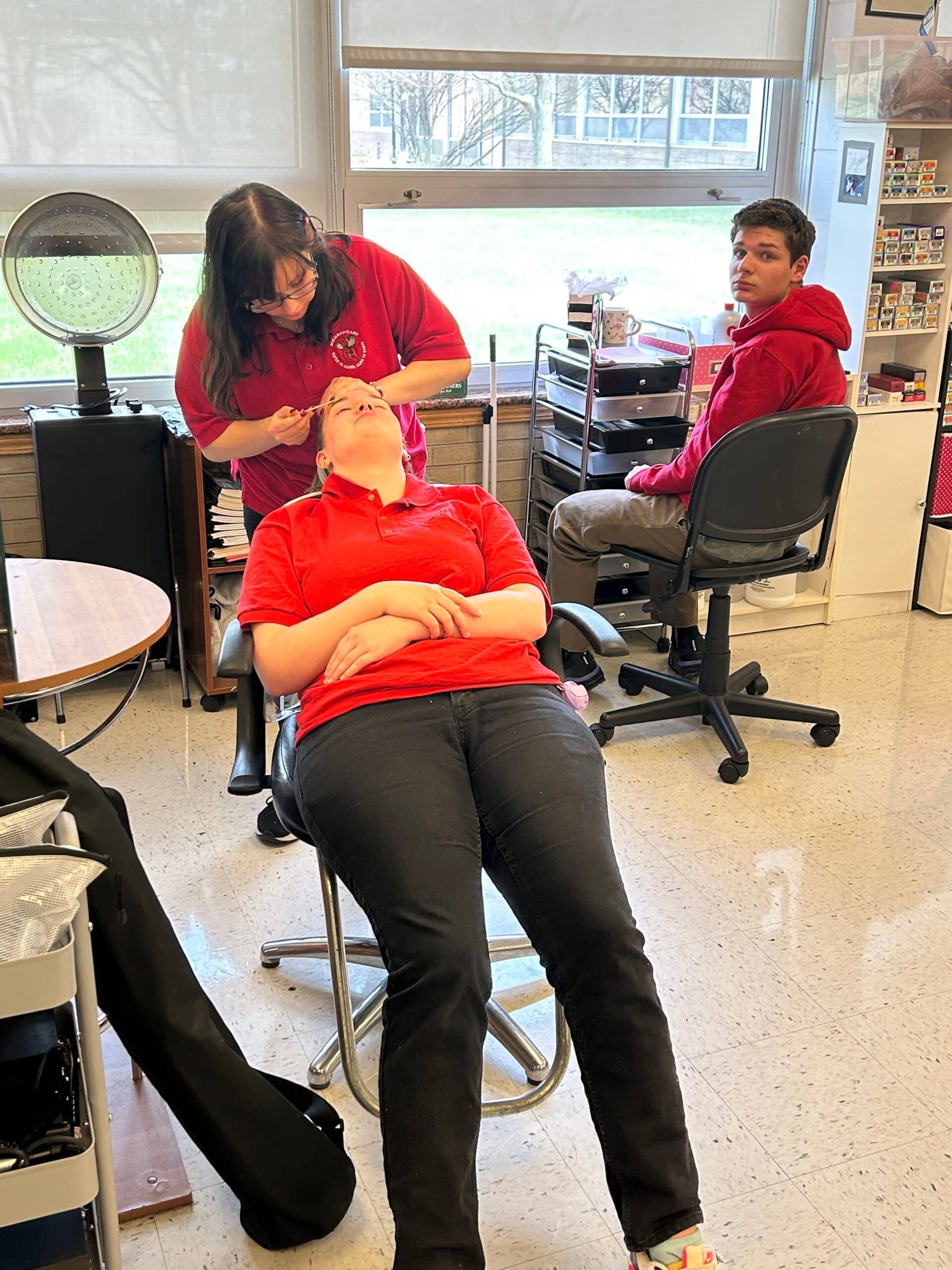 One woman sits on a chair, with her head reclined back. Standing next to her is another woman, who looks at her eyebrows with a waxing kit in her hand.