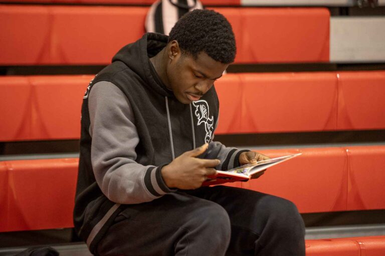 A young man reading a yearbook while seated on a bleacher in a peaceful park environment.